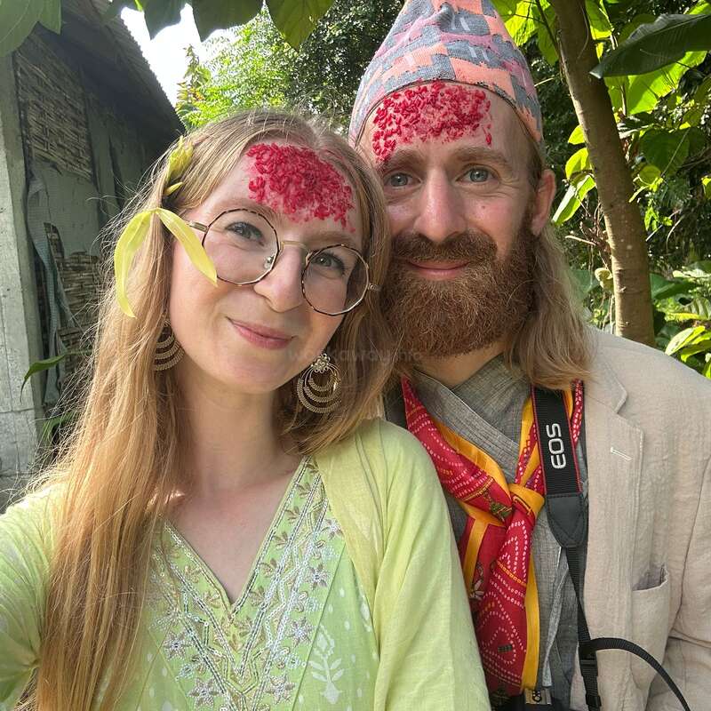 A smiling couple with tika on their foreheads pose outdoors. Both have traditional accessories, surrounded by lush greenery, enjoying a festive or cultural celebration together.