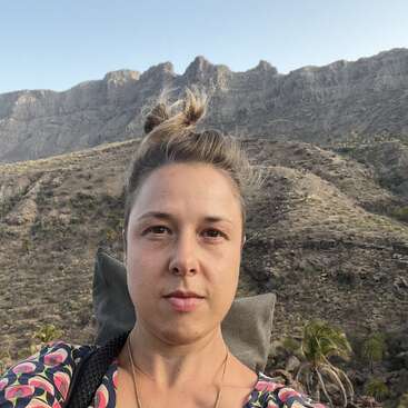 Uma mulher tira uma selfie ao ar livre, com uma paisagem montanhosa e rochosa ao fundo. Ela tem cabelos claros presos em um coque, veste uma blusa estampada e parece tranquila.