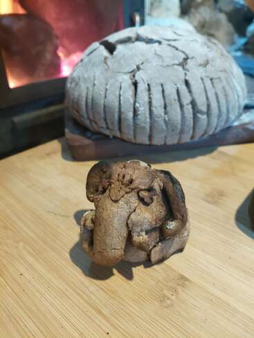 Um pão rústico está sobre uma tábua de madeira. À frente, está exposta uma pequena figura esculpida em pão que lembra um mamute lanudo, com um fundo aconchegante.