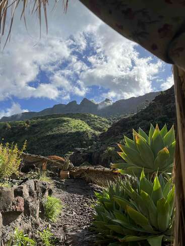 Montanhas verdejantes sob um céu dramático repleto de nuvens, agaves em primeiro plano, um caminho rochoso e um telhado de palha emoldurando parcialmente a bela paisagem natural.