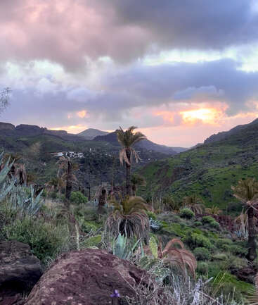 Um céu dramático ao pôr do sol, com nuvens cor-de-rosa, paira sobre colinas verdes, palmeiras, cactos e rochas. Montanhas distantes e uma pequena vila completam a paisagem tranquila.