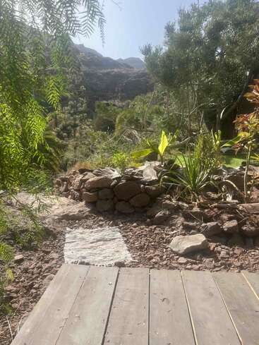 Uma cena de jardim banhada pelo sol mostra um deck rústico de madeira, um pequeno tapete tecido, uma parede de pedra, vegetação exuberante e montanhas ao fundo, sob um céu limpo.