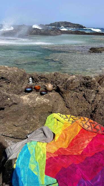 Uma cena animada na praia mostra uma toalha colorida sobre as rochas, um pequeno serviço de chá ao lado, com as ondas do mar batendo ao fundo num dia ensolarado e claro.