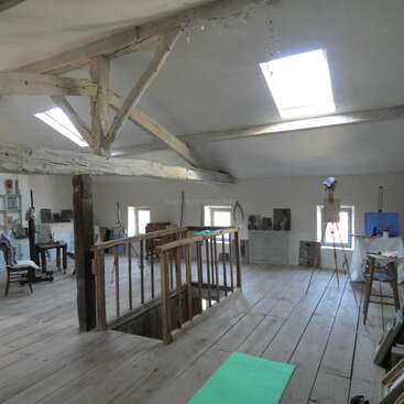 This image depicts a spacious, rustic art studio with wooden floors, exposed beams, skylights, easels, paintings, and art supplies, creating a creative and airy atmosphere.
