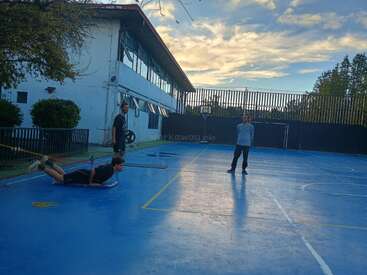 Three people are on a blue outdoor sports court. One person is being pulled on a board with a rope, while two others observe and smile nearby.