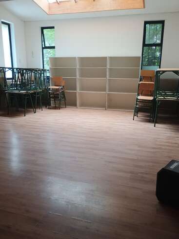This image shows an empty classroom with wooden floors, stacked chairs, empty bookshelves, and large windows letting in natural light. The space looks clean and organized.