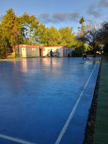 A blue outdoor basketball court reflecting sunlight, surrounded by trees and bushes. Portable cabins and a few people can be seen in the background, enjoying the scene.