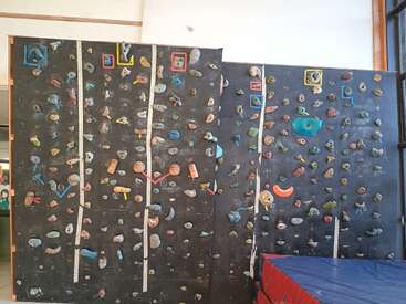 The image shows an indoor bouldering climbing wall with various colorful handholds and footholds. There are white tape lines dividing climbing routes and safety mats below.