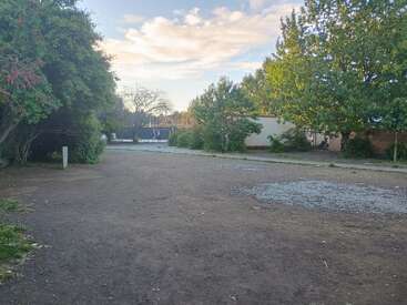 A dirt pathway is surrounded by green trees and bushes, with buildings in the background. The sky is partly cloudy, with soft evening sunlight illuminating the scene.