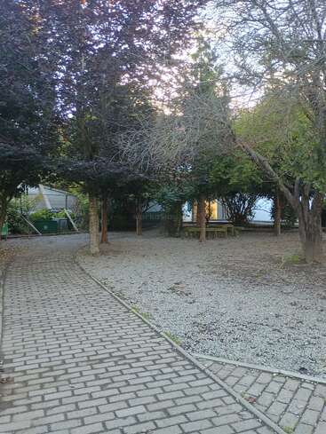A paved walkway curves through a quiet garden with leafy trees and gravel ground. Benches and a table sit under the shade, bathed in soft sunlight.