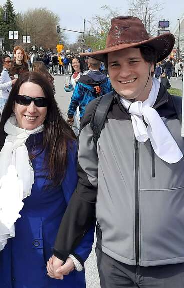 A smiling man and woman hold hands at an outdoor event, surrounded by a crowd. The man wears a cowboy hat, both wear white scarves, and jackets.