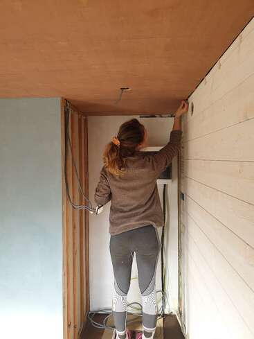 A woman stands on the floor, working on wiring or installing something near the ceiling inside a partially constructed or renovated room, wearing casual athletic clothing and sneakers.