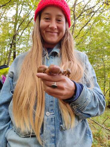 A woman with long blonde hair, wearing a red knit hat and denim jacket, smiles while holding mushrooms in a forest with lush green trees.