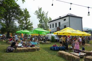 People are gathered at an outdoor festival, sitting on hay bales under green and yellow umbrellas. There are trees, a white building, and string lights overhead.