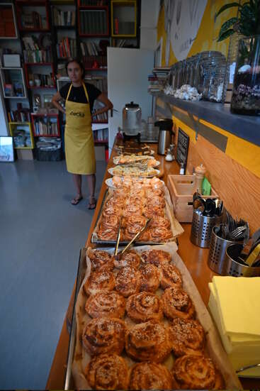 A woman in a yellow apron stands by a long table filled with pastries and snacks, inside a cozy bookstore café with bookshelves and jars.