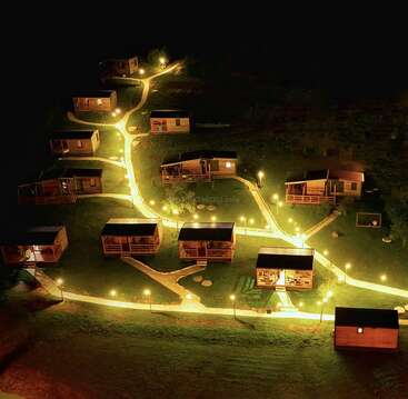 Aerial night view of cozy wooden cabins arranged along illuminated winding pathways, surrounded by darkness, creating a warm and inviting atmosphere in a peaceful rural setting.
