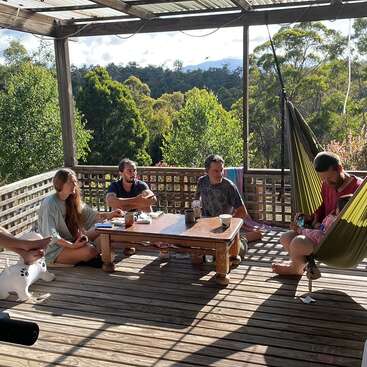 The image depicts a serene outdoor setting, featuring a wooden deck with a table and hammock, surrounded by lush trees and a mountainous backdrop on a sunny day.