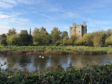 A tranquil river with two swans flows before lush greenery. In the background, a historic stone castle rises against a blue sky, creating a peaceful scene.