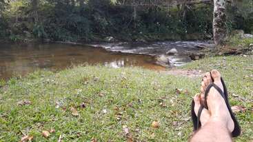 A person relaxes on grassy riverbank, wearing flip-flops, with feet stretched out toward a calm river surrounded by lush green forest and tranquil nature.