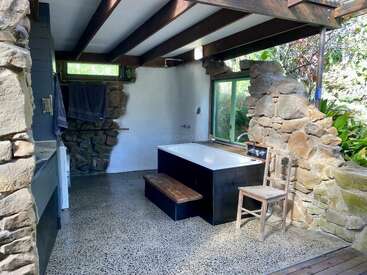 Dieses rustikale Badezimmer hat Steinwände, freiliegende Holzbalken und große Fenster mit üppigem Grün draußen. Eine Badewanne, ein Holzstuhl und Handtücher machen den gemütlichen Raum komplett.