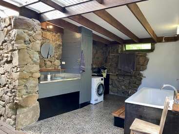 Dieses rustikale Badezimmer hat Steinwände, freiliegende Holzbalken, eine Badewanne, eine Waschmaschine, einen runden Spiegel und moderne Armaturen. Oberlichter und natürliches Licht machen die Atmosphäre noch gemütlicher.