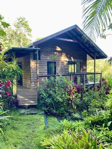 Eine hübsche Holzhütte liegt eingebettet in üppigem, grünem tropischem Laub. Blühende Pflanzen und leuchtendes Grün umgeben die gemütliche Veranda und schaffen eine ruhige, einladende Atmosphäre.