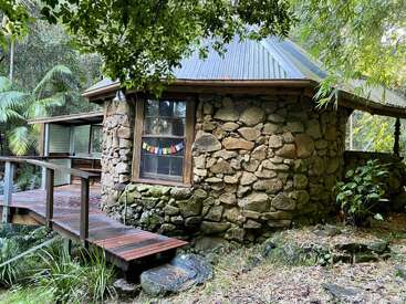 Auf dem Bild sieht man eine kleine runde Steinhütte mit Blechdach, umgeben von üppig grünen Bäumen. Ein Holzsteg führt zum Eingang. Eine echt ruhige Umgebung.
