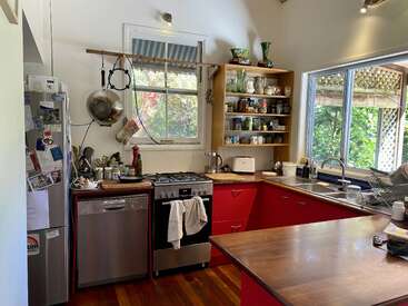 Helle, gemütliche Küche mit roten Schränken, Holzarbeitsplatten, offenen Regalen, großen Fenstern, hängenden Töpfen, Spülmaschine, Kühlschrank voller Magnete und auf den Arbeitsplatten verstreuten Küchenutensilien. Einladende Atmosphäre.