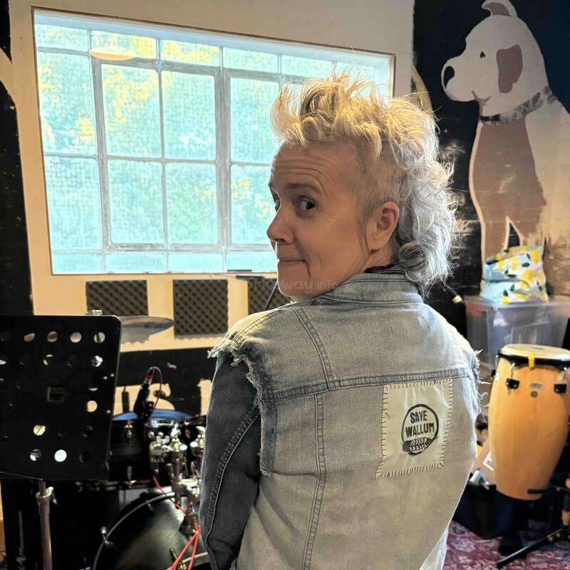 Eine Person mit Irokesenfrisur, die eine Jeansweste mit "Save Wallum"-Aufnäher trägt, steht in einem Musikstudio mit Schlagzeug, einer Wandmalerei und Instrumenten.
