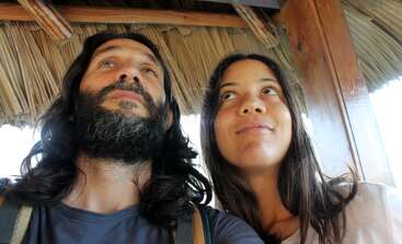 A man and a woman with long hair are looking up, standing closely together under a thatched roof, appearing relaxed and thoughtful in the natural light.