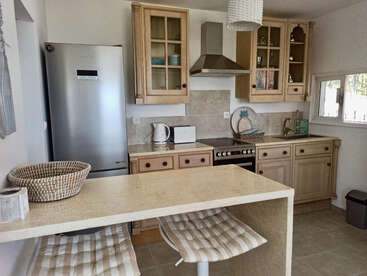 This cozy kitchen features wooden cabinets, stainless steel appliances, a marble island with two striped stools, a wicker basket, and bright natural light from a small window.