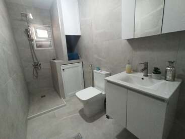 This image shows a modern, compact bathroom with light gray tiles, a walk-in shower, toilet, vanity sink, mirror cabinets, and minimalistic decor, including soap and a small plant.