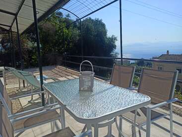 A sunlit outdoor patio features a glass table with chairs, a lantern centerpiece, overlooking a scenic view of the sea, mountains, and houses below.