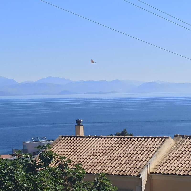 The image depicts a serene scene of a house with a tiled roof, overlooking a vast body of water and distant mountains under a clear blue sky.