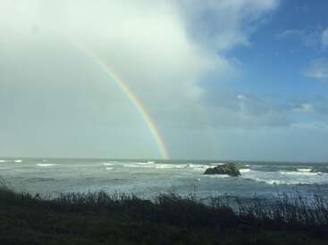 Um arco-íris vibrante se estende por um céu nublado acima do oceano, ondas batem contra uma rocha solitária, enquanto a grama selvagem se alinha na costa ventosa e tempestuosa.