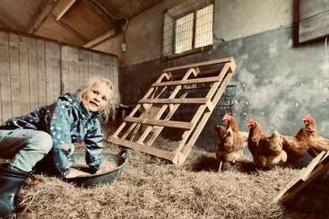 Ein kleines Kind in einem blauen Regenmantel sitzt in einer Scheune und spielt mit einer Schüssel, während vier Hühner in der Nähe auf dem strohbedeckten Boden stehen. Gemütliche Atmosphäre.