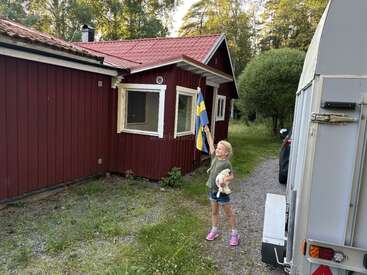 Ein junges Mädchen steht vor einem roten Holzhaus und hält eine schwedische Flagge und ein Stofftier in der Hand. Im Hintergrund sind ein Wohnwagen und Bäume zu sehen.
