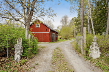 Eine charmante rote Scheune steht neben einem Feldweg, umgeben von üppigen grünen Bäumen, Holzzäunen und Steinmarkierungen in einer friedlichen ländlichen Umgebung.