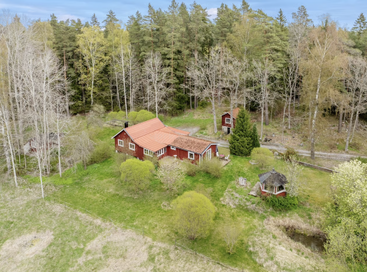 Ein charmantes rotes Häuschen mit Nebengebäuden liegt inmitten von üppigem Grün und Bäumen, umgeben von einem Wald. Friedliche ländliche Umgebung, Pavillon und ein gewundener Feldweg.