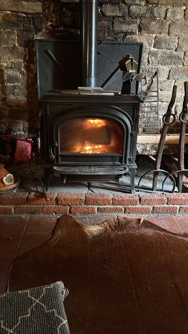 Un poêle à bois accueillant est adossé à un mur de pierre rustique, diffusant une chaleur réconfortante. Les outils et accessoires pour le feu sont posés à proximité. Le foyer en briques et le tapis en peau d'animal ajoutent au charme de la pièce.