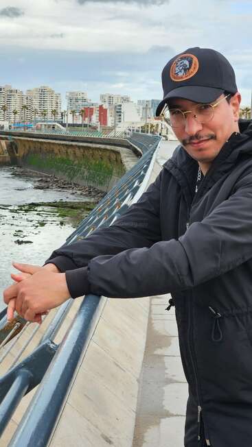Un homme vêtu d'une veste noire et d'une casquette est appuyé contre la balustrade d'un pont, avec en arrière-plan les bâtiments de la ville, l'eau et un ciel nuageux. Il sourit légèrement.