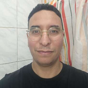Jemand mit kurzen Locken und Brille lächelt leicht. Die Person trägt ein schwarzes Shirt und steht vor einer gefliesten Wand und einem bunten, gestreiften Duschvorhang.