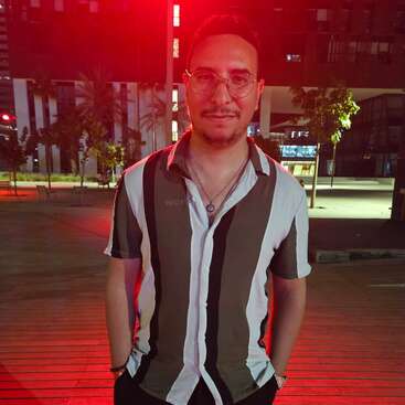 A man wearing glasses and a vertically striped shirt stands outdoors at night, illuminated by red lights. Trees and modern buildings are visible in the background.