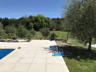The image depicts a serene backyard scene featuring a pool, lush greenery, and a statue, set against a clear blue sky on a sunny day.