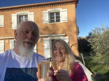 The image depicts a man and woman toasting with champagne glasses in front of a house, set against a serene backdrop of a clear blue sky and lush greenery.