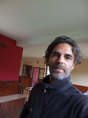 Un homme aux cheveux mi-longs et à la barbe prend un selfie à l'intérieur. Il porte une veste noire ; la pièce a des murs et des meubles rouges et en bois.