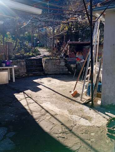Une cour ensoleillée avec un sol en pierre, un balai, une échelle, un four extérieur, du bois de chauffage empilé, des escaliers, des vignes en surplomb, une table et un jardin, créant une atmosphère rustique et paisible.
