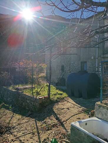 Les rayons du soleil éclairent une maison rustique en pierre et une cour envahie par la végétation. Des branches d'arbres dénudées, une citerne noire, une vieille baignoire et des outils de jardinage créent une scène rurale et tranquille.