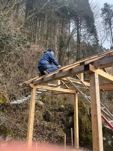 Eine Person in einer blauen Jacke baut eine Holzkonstruktion im Freien, kniet auf dem Dach, umgeben von Wald und Bäumen, und benutzt eine Leiter als Zugang.