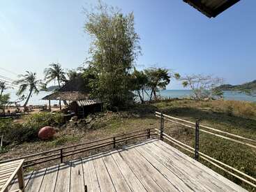 Une terrasse en bois surplombe une pelouse, des arbres tropicaux et une hutte au toit de chaume près de la plage. Les palmiers et l'eau bleue de l'océan complètent ce paysage serein.